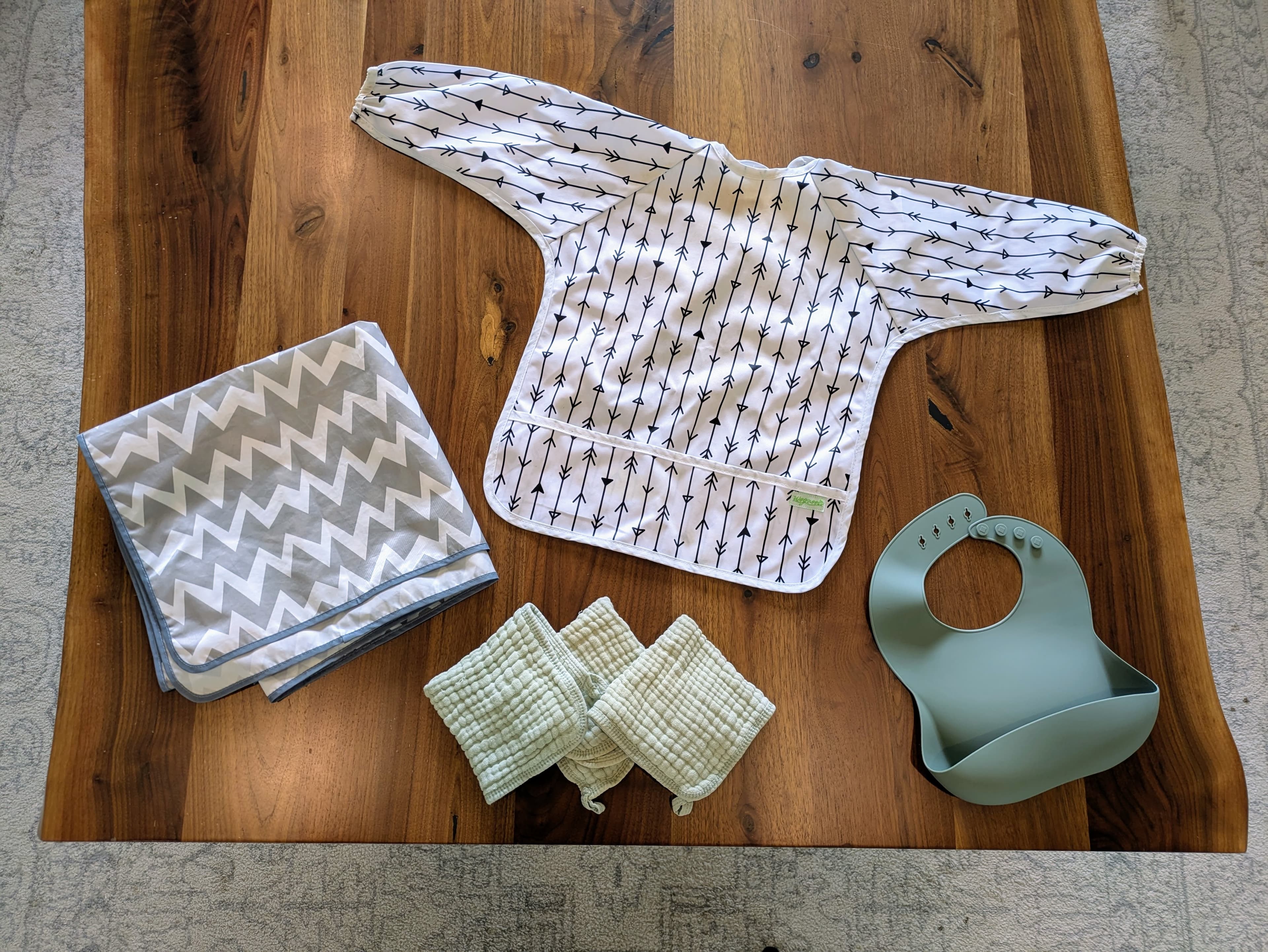 A photograph of a wooden table with a bib, a smock, a splat mat, and some cloths