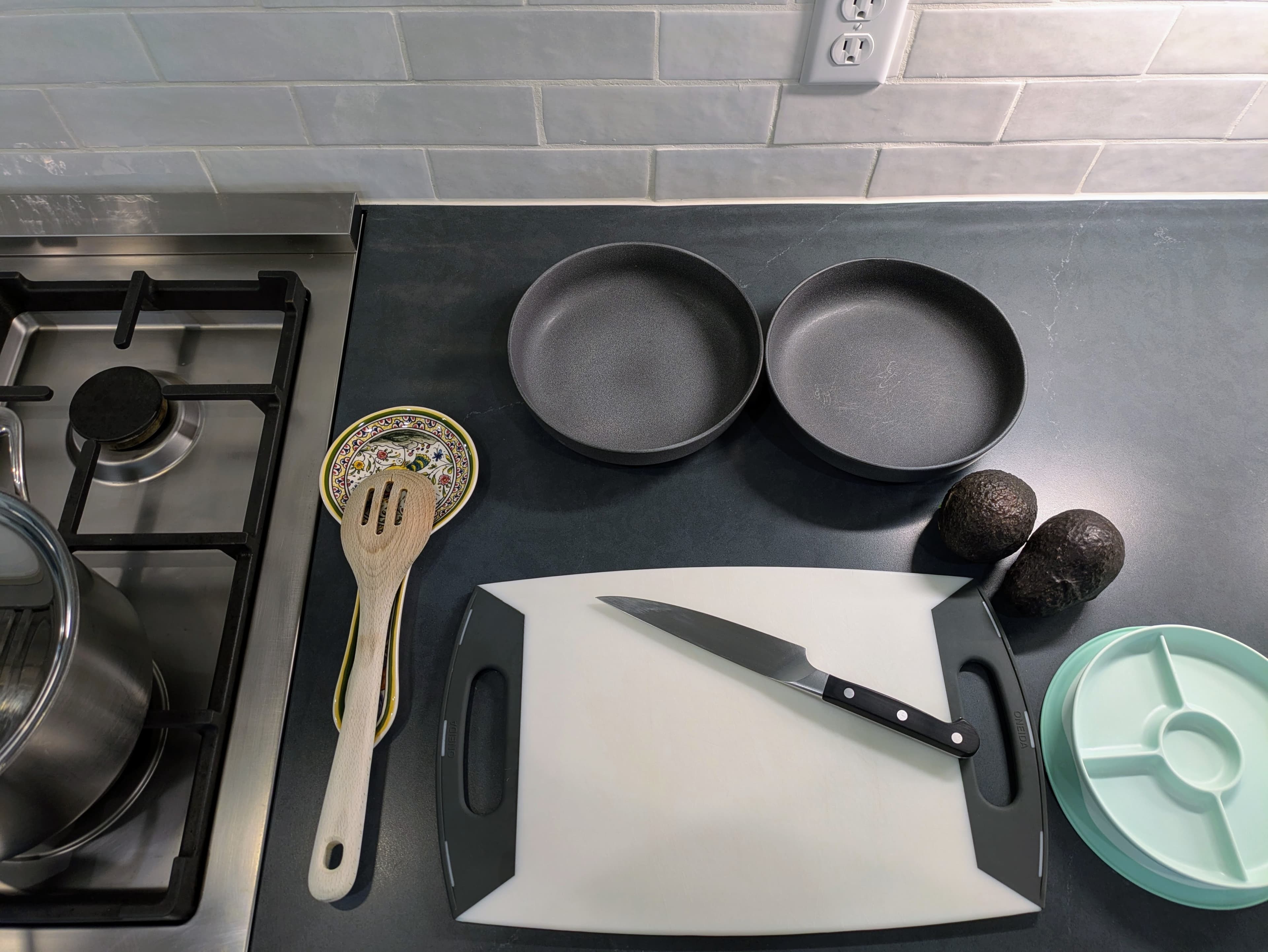 A photograph of a dark countertop with a cutting board, a knife, two adult bowls, a child's plate, two avocados, and a spoon on a spoonrest
